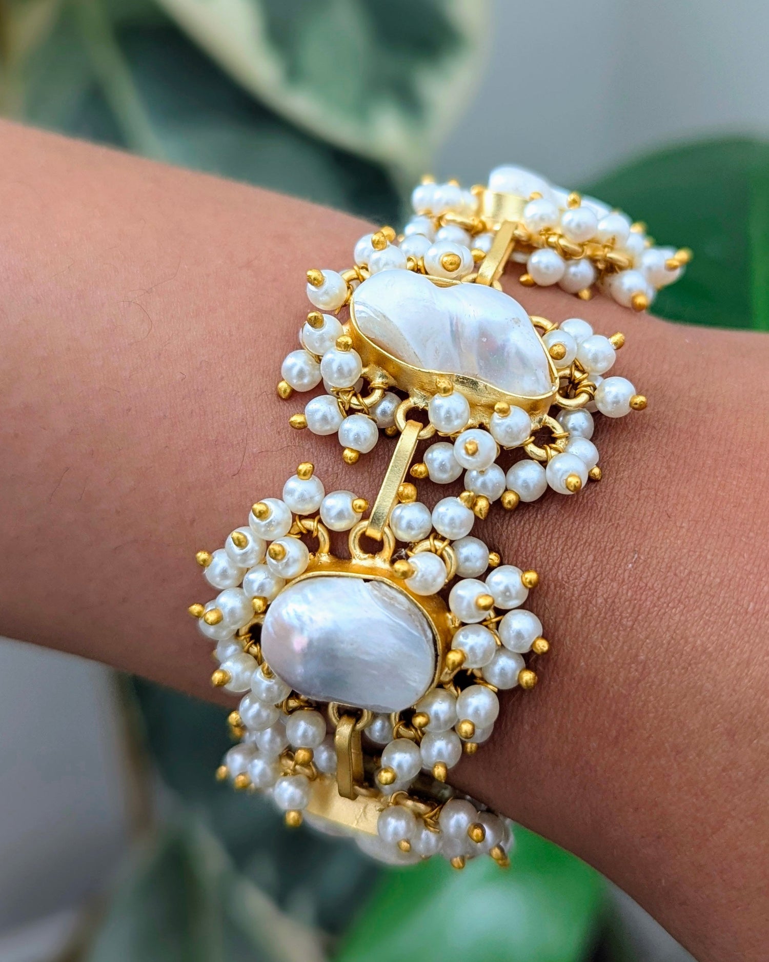 Gold and pearl bracelet on a wrist with a blurred green plant background