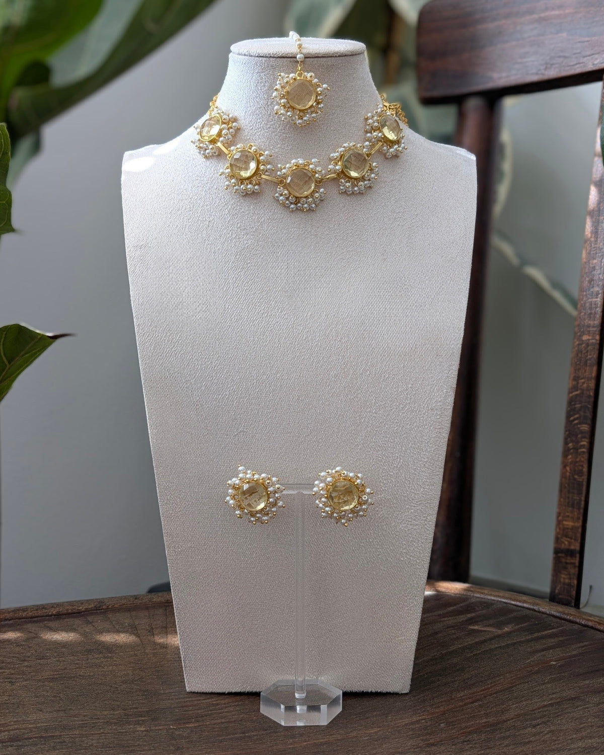 Gold and white floral jewelry set displayed on a white mannequin bust, including a necklace, matching earrings, and a small pendant, set on a wooden table with green plants in the background.