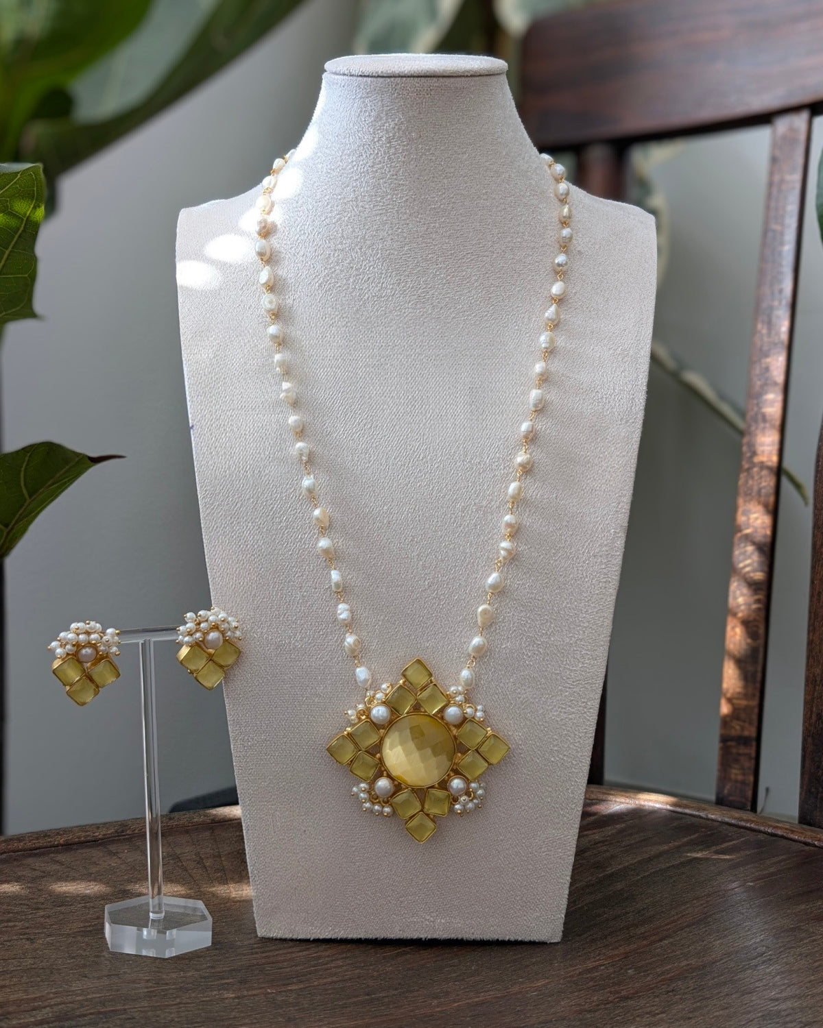 A gold and pearl statement necklace displayed on a cream fabric mannequin bust, with matching gold and pearl stud earrings on a clear acrylic stand beside it.