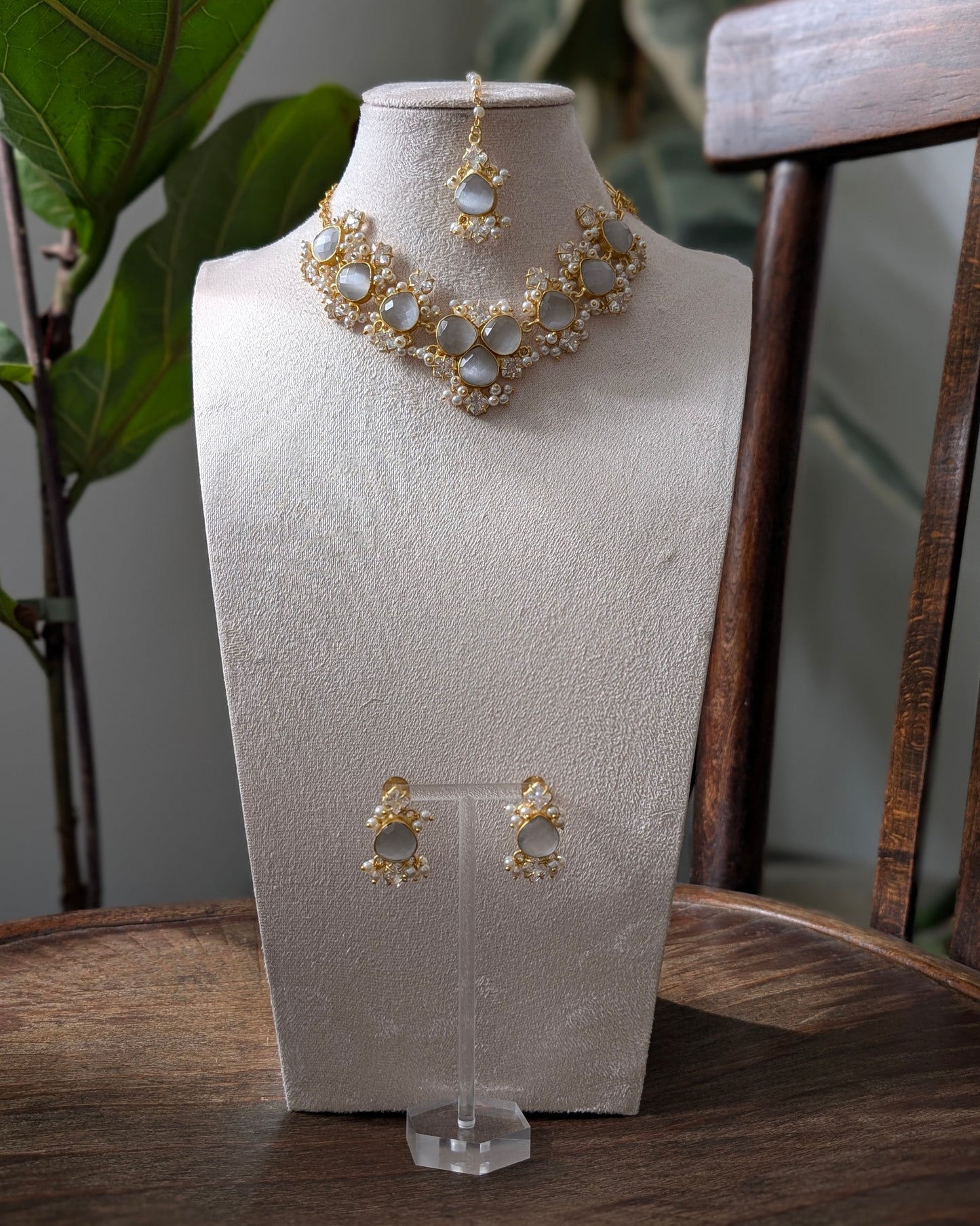 Gold-toned jewelry set displayed on a textured cream jewelry bust, including a statement necklace, matching pendant, and earrings, set on a wooden table with a chair and green leaves in the background.