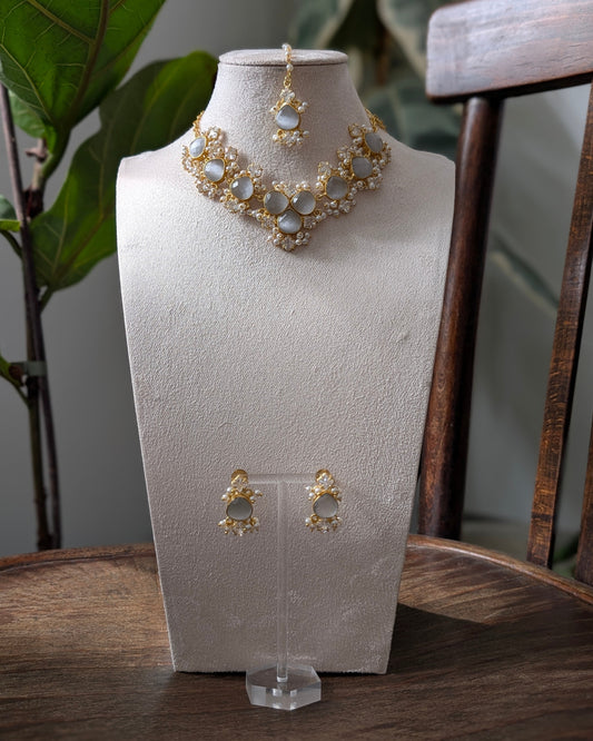 Gold-toned jewelry set displayed on a textured cream jewelry bust, including a statement necklace, matching pendant, and earrings, set on a wooden table with a chair and green leaves in the background.