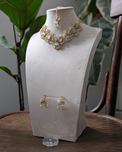 Gold and white stone jewelry set including a necklace, matching earrings, and a maang tikka, displayed on a textured white jewelry bust on a wooden table with a plant in the background.