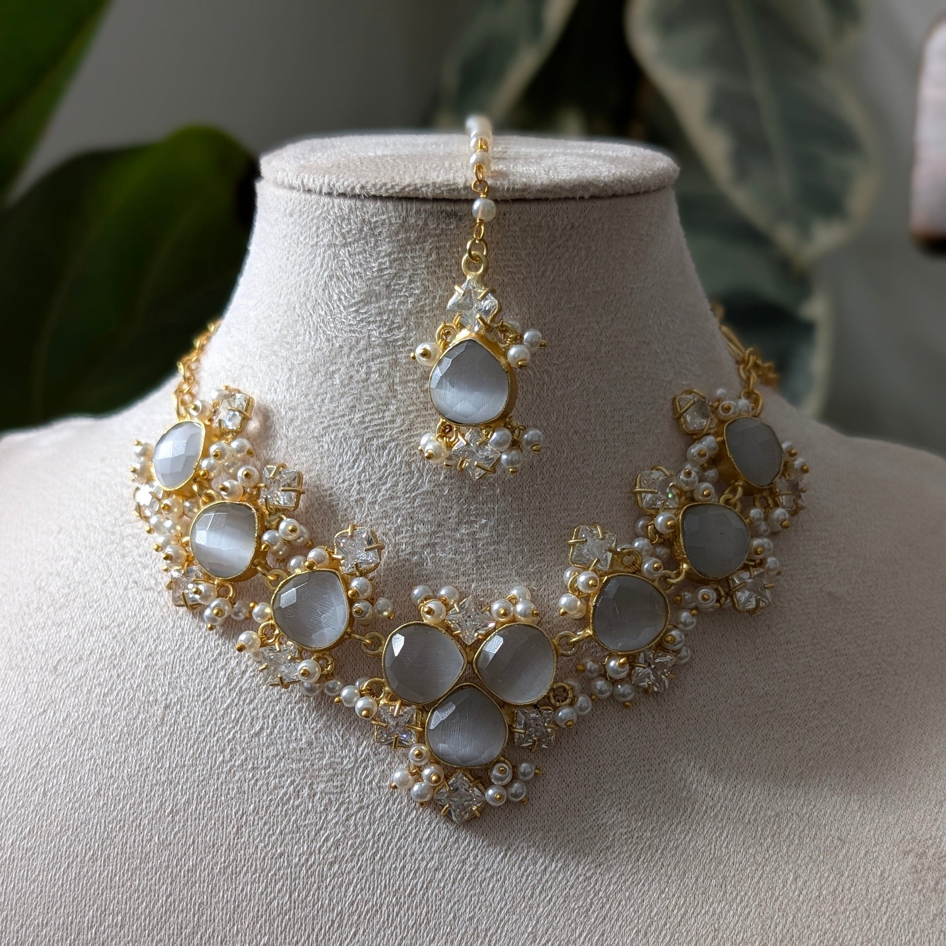 Gold and pearl jewelry set featuring a choker necklace and matching maang tikka, displayed on a cream-colored mannequin bust with green leaves in the background.