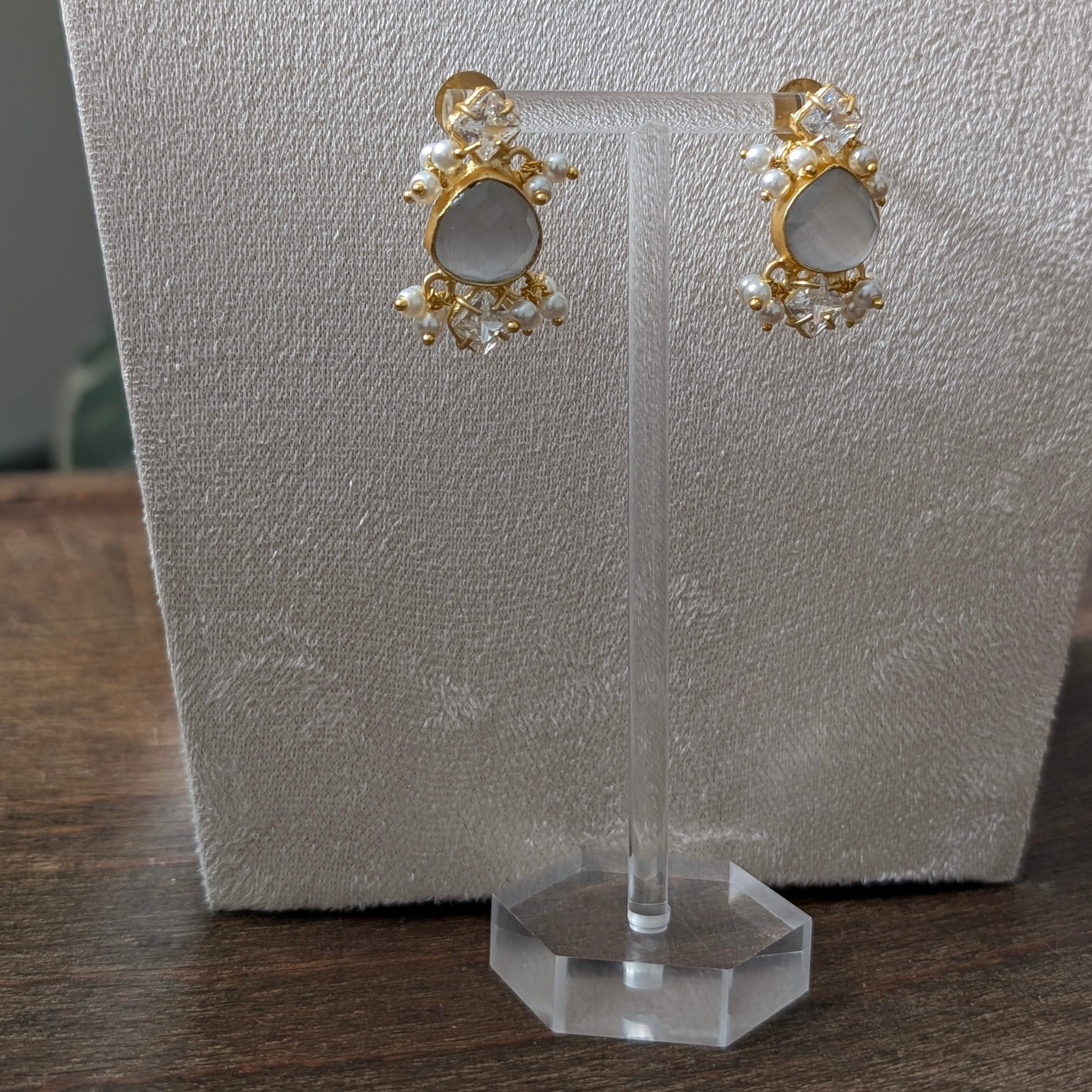 A pair of gold-toned statement earrings with oval white stones and small pearl-like accents, displayed on a clear stand against a textured white card on a wooden surface.