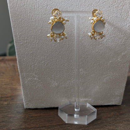 A pair of gold-toned statement earrings with oval white stones and small pearl-like accents, displayed on a clear stand against a textured white card on a wooden surface.