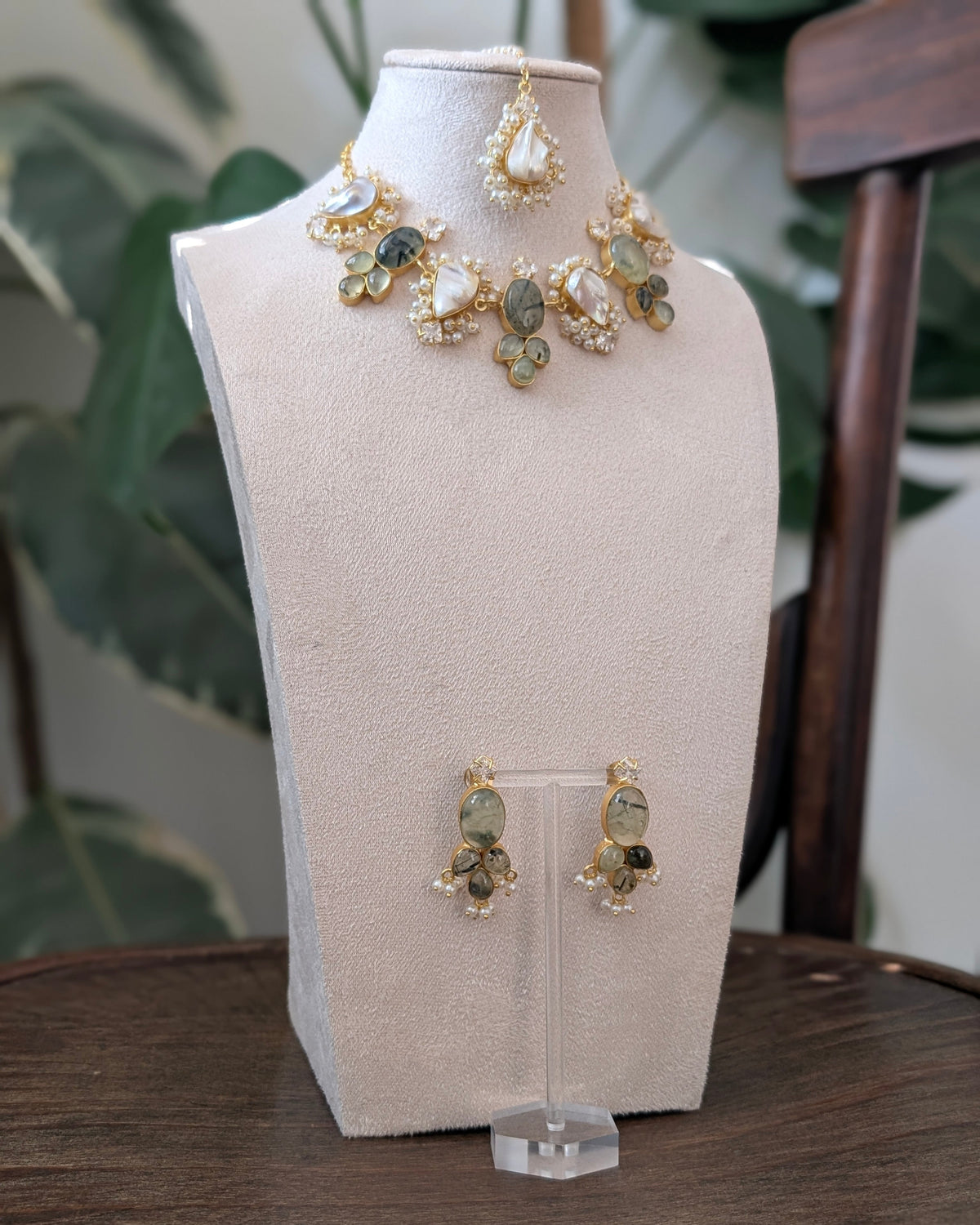 Gold and green stone jewelry set displayed on a beige jewelry bust, including a necklace and matching earrings, with a wooden table and green plants in the background.