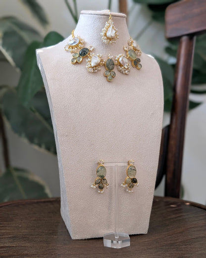 Gold and green stone jewelry set displayed on a beige jewelry bust, including a necklace and matching earrings, with a wooden table and green plants in the background.