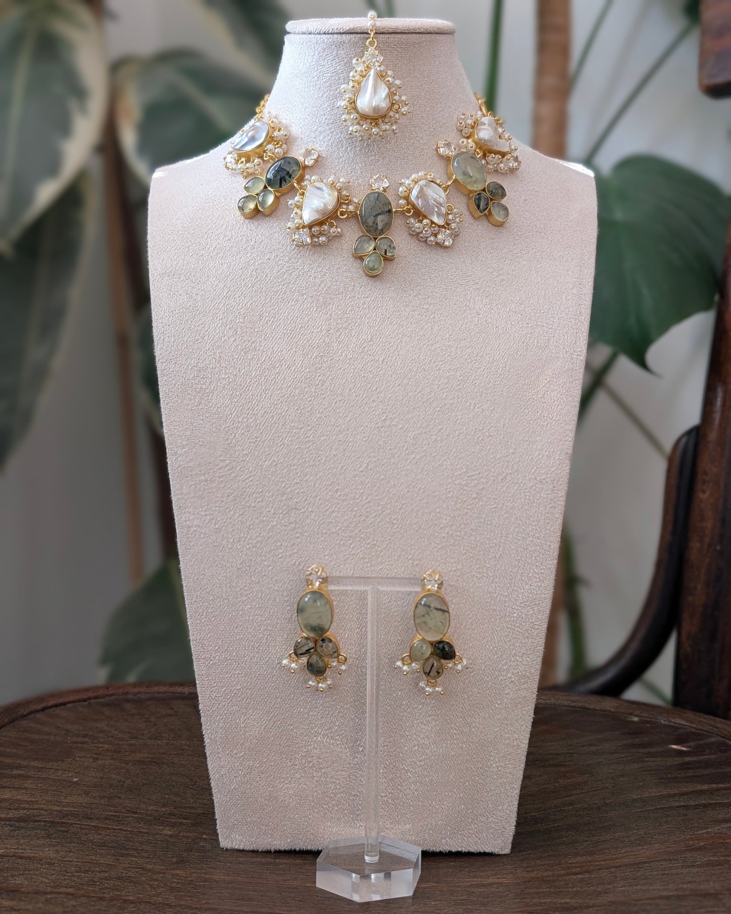 Gold-toned jewelry set displayed on a cream jewelry bust, including a statement necklace, matching earrings, and a top piece, with a blurred indoor plant and wooden table in the background.