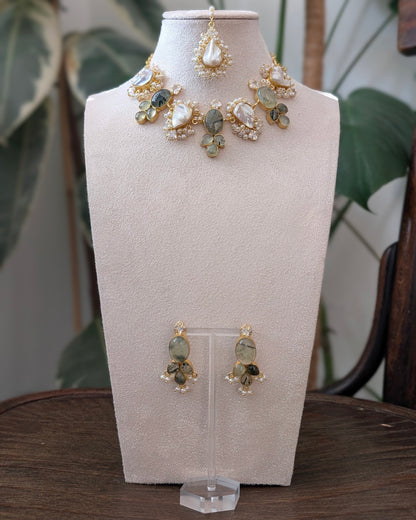 Gold-toned jewelry set displayed on a cream jewelry bust, including a statement necklace, matching earrings, and a top piece, with a blurred indoor plant and wooden table in the background.