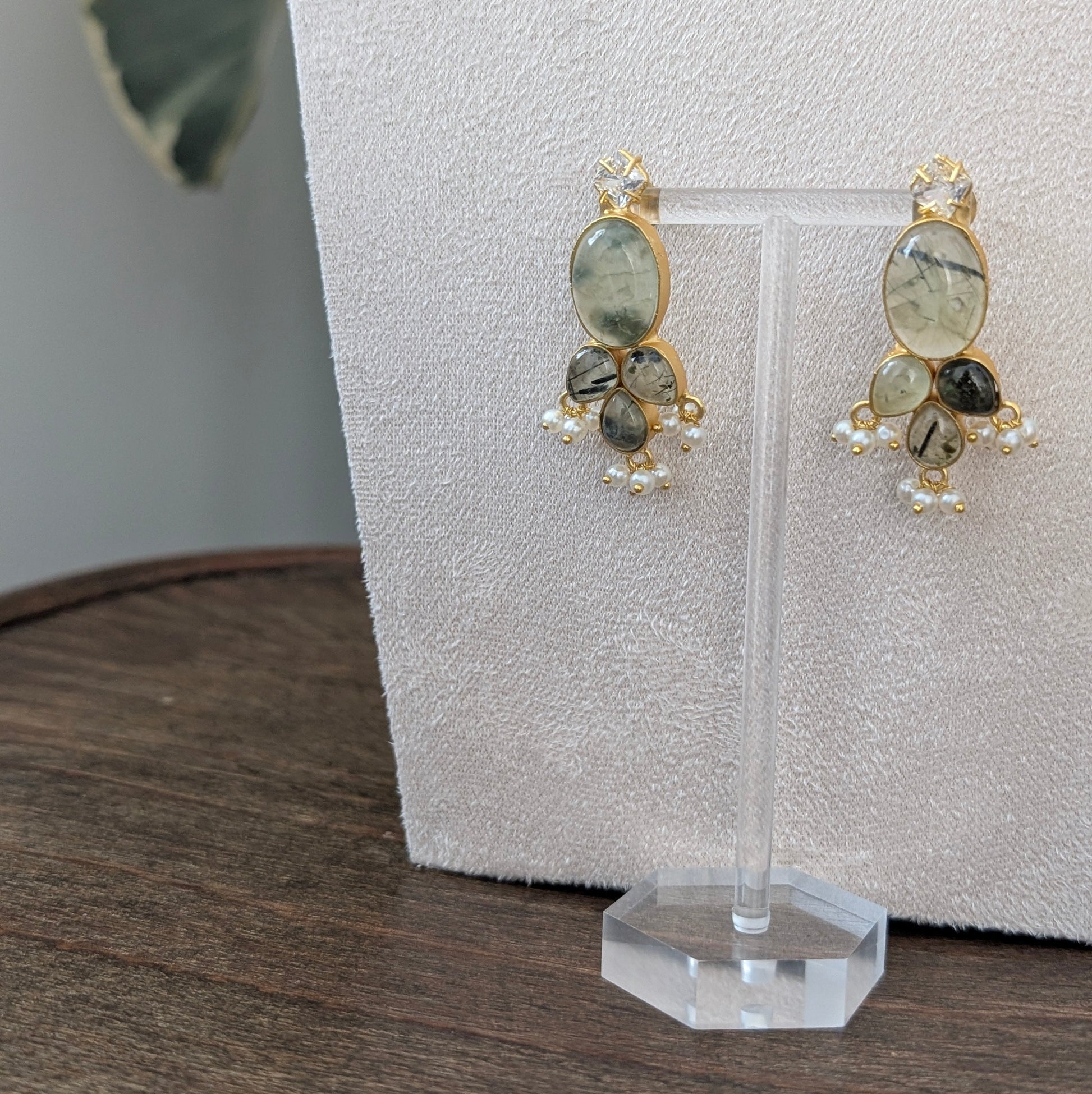 A pair of gold-toned dangle earrings with large oval greenish-gray stones and smaller dark and pearl accents, displayed on a clear stand against a textured light backdrop.