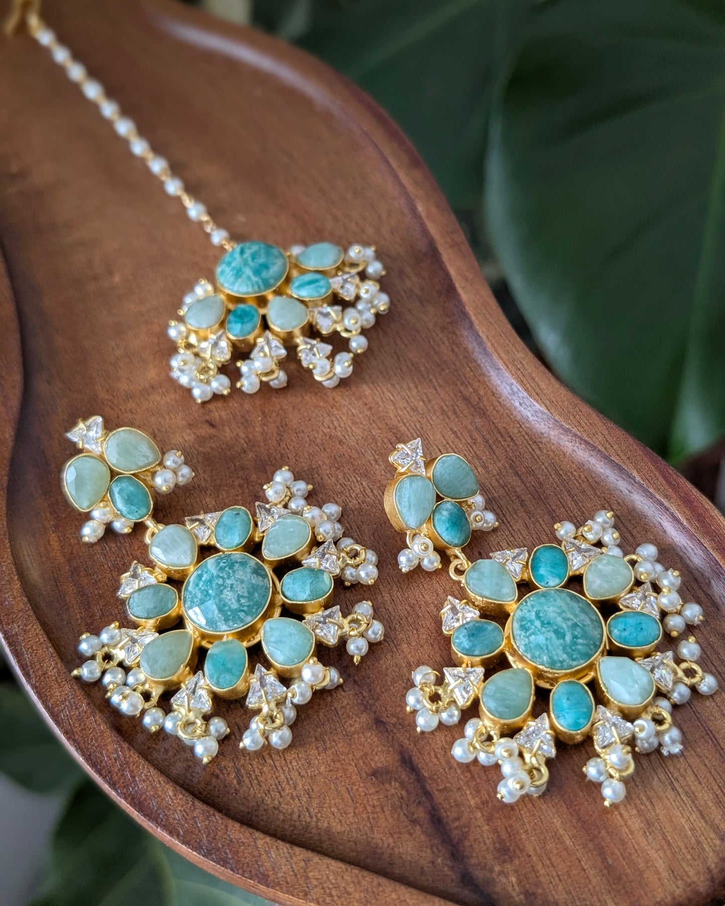 Gold-toned traditional Indian jewelry set featuring a maang tikka and matching earrings, embellished with light blue-green stones and small white pearls, displayed on a wooden tray with green leaves in the background.