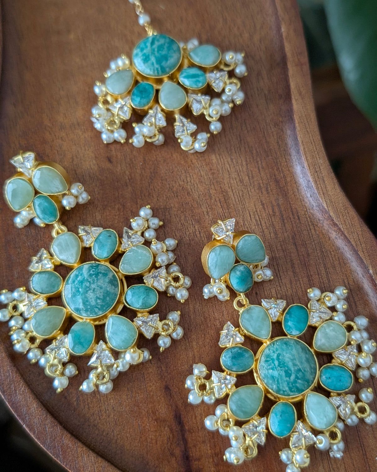 A gold-plated jewelry set featuring a necklace and two matching earrings with round turquoise-blue stones and small white pearl-like accents, arranged on a curved wooden surface.