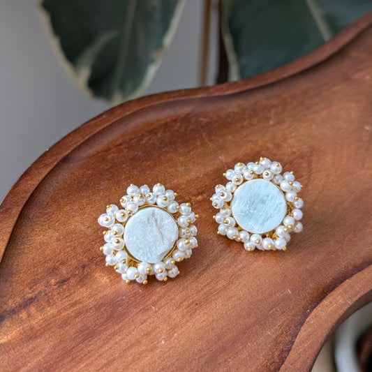A pair of gold-toned stud earrings with round pale blue centers surrounded by small white pearl-like beads, displayed on a curved wooden surface with blurred green leaves in the background.