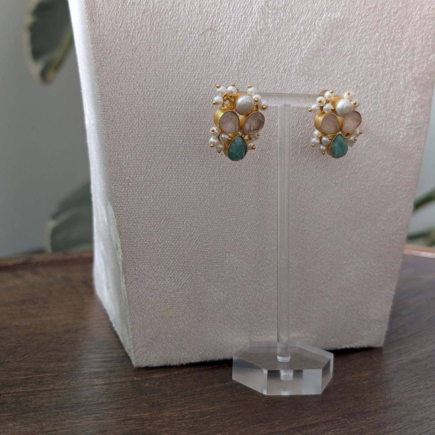 A pair of gold and pearl stud earrings with small green stone accents, displayed on a clear acrylic stand against a textured white jewelry bust.