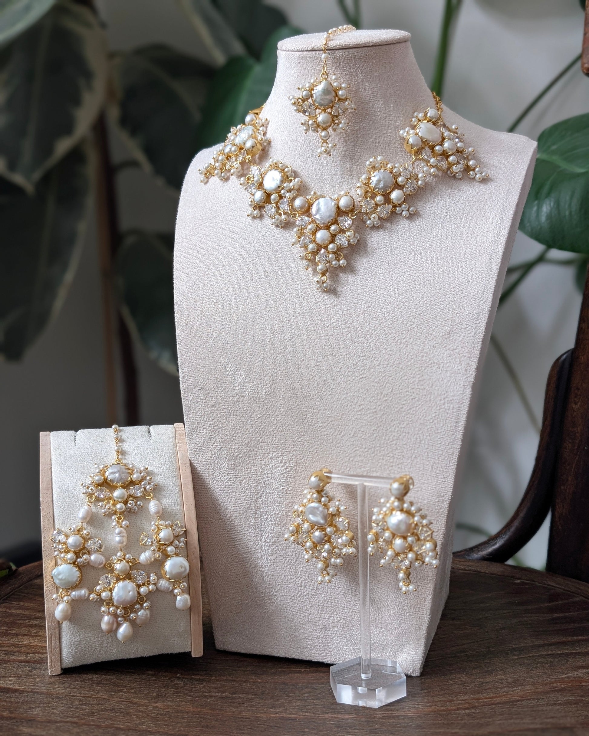 A jewelry set displayed on a cream fabric mannequin bust and small stands, featuring a necklace, matching earrings, and a pair of drop earrings with cream faux pearls and gold-toned metal details, set against a background of indoor plants and a wooden table.