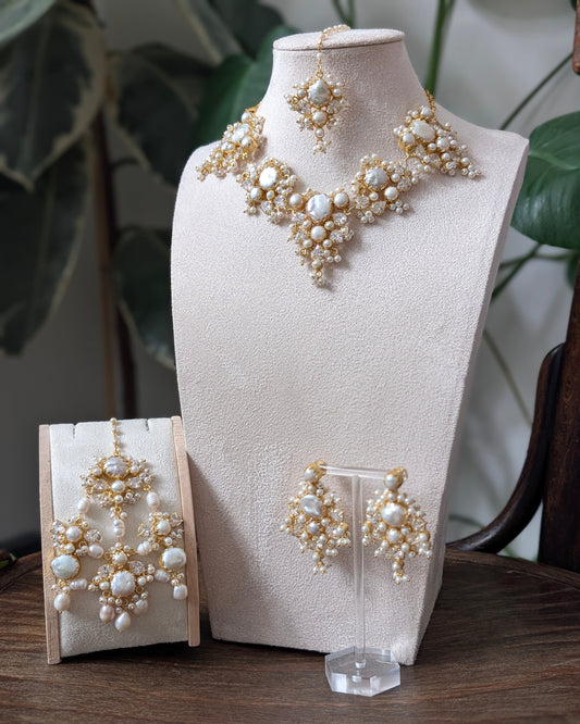 A jewelry set displayed on a cream fabric mannequin bust and small stands, featuring a necklace, matching earrings, and a pair of drop earrings with cream faux pearls and gold-toned metal details, set against a background of indoor plants and a wooden table.