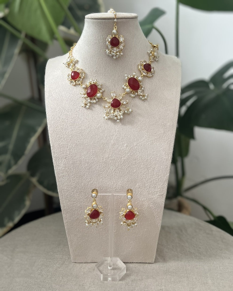 Red ruby and pearls indian jewelry set with necklace and earrings on a white stand with green leaves in the background