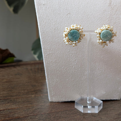 A pair of bohemian-style earrings with round light blue turquoise-like centers surrounded by white pearl-like beads and small white star-shaped accents, displayed on a clear acrylic stand against a textured white backdrop.