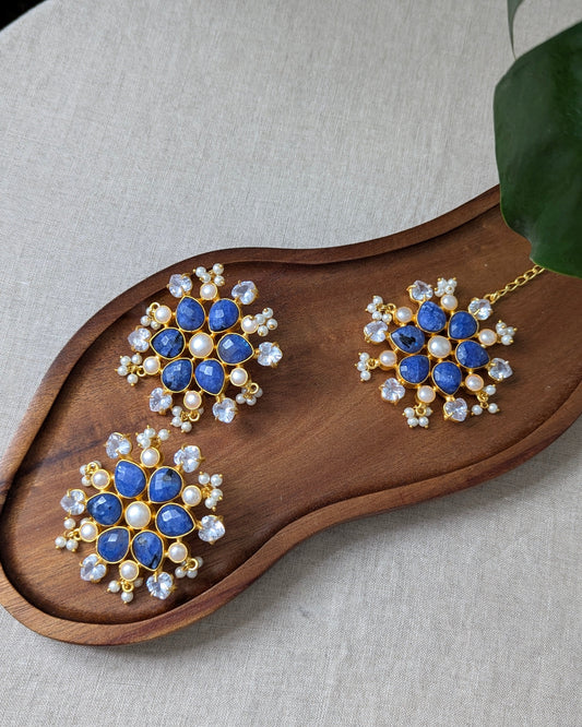 Pair of earrings with blue gemstones and pearl-like stones on a wooden tray.