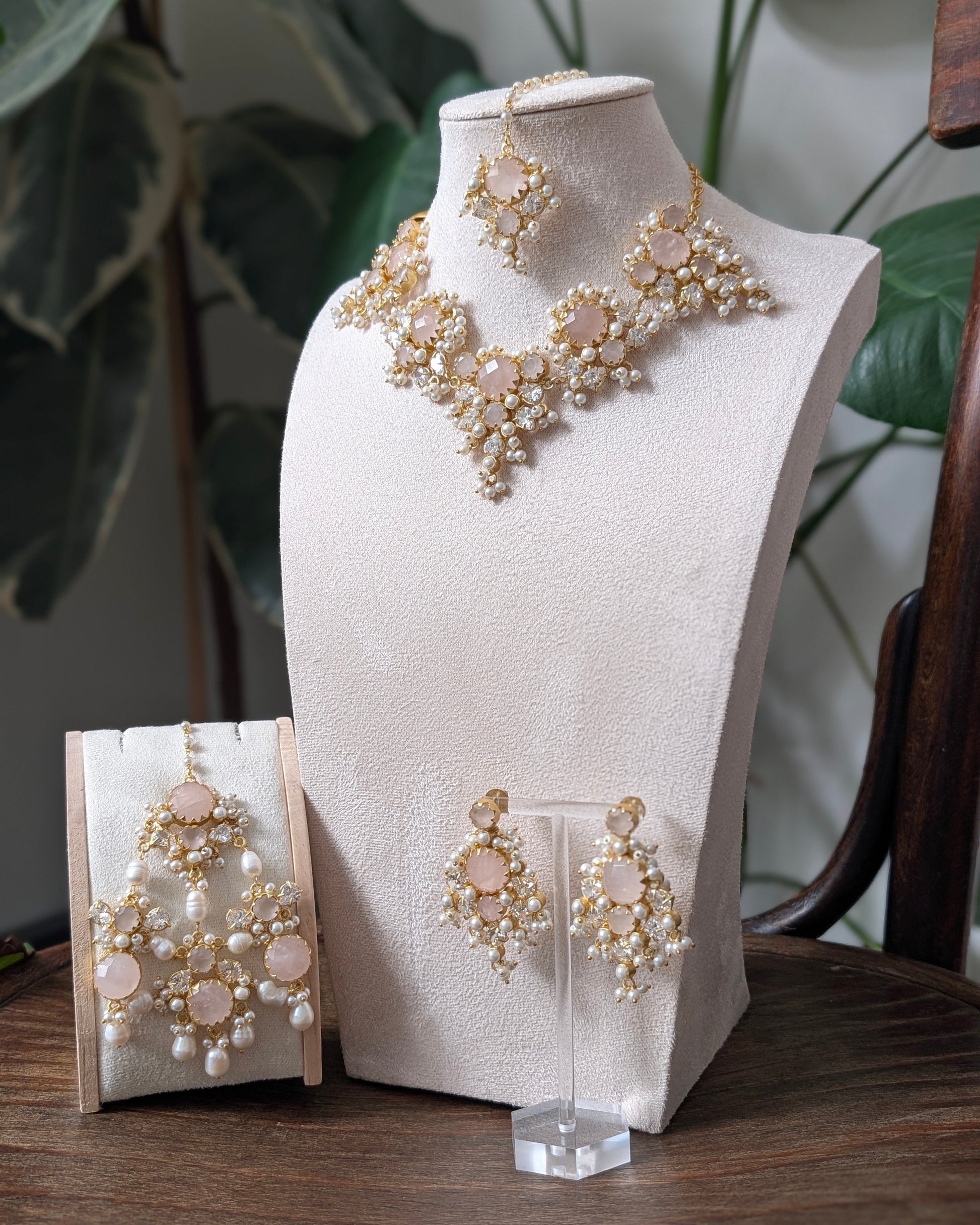 A gold and pearl bridal jewelry set displayed on a cream-colored mannequin bust and a small stand, featuring a statement necklace, matching earrings, and a pair of drop earrings with pink stones, set against a background of indoor plants.
