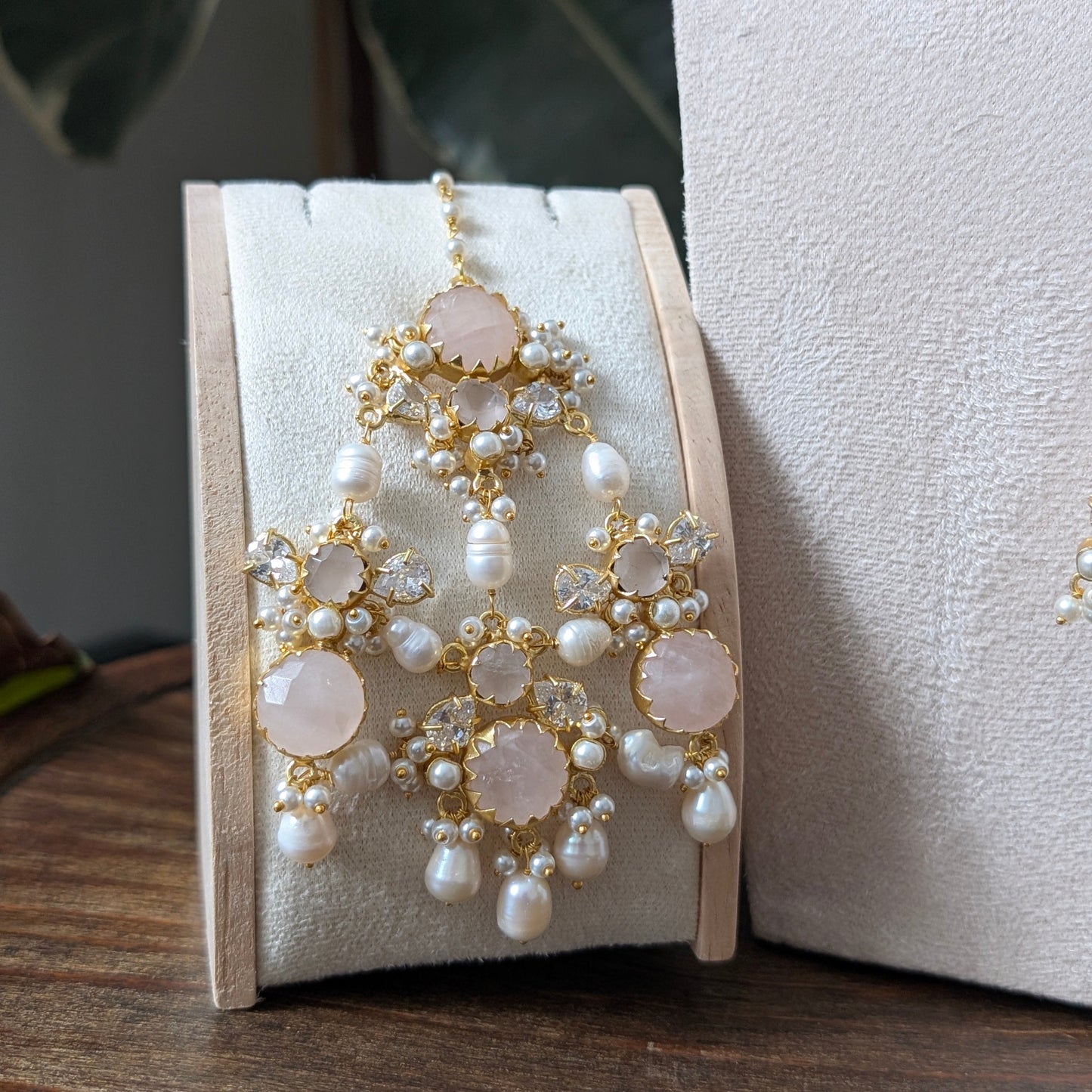 A pair of ornate gold-toned jhumar with white pearls and pale pink stone accents, displayed on a cream jewelry stand on a wooden surface.