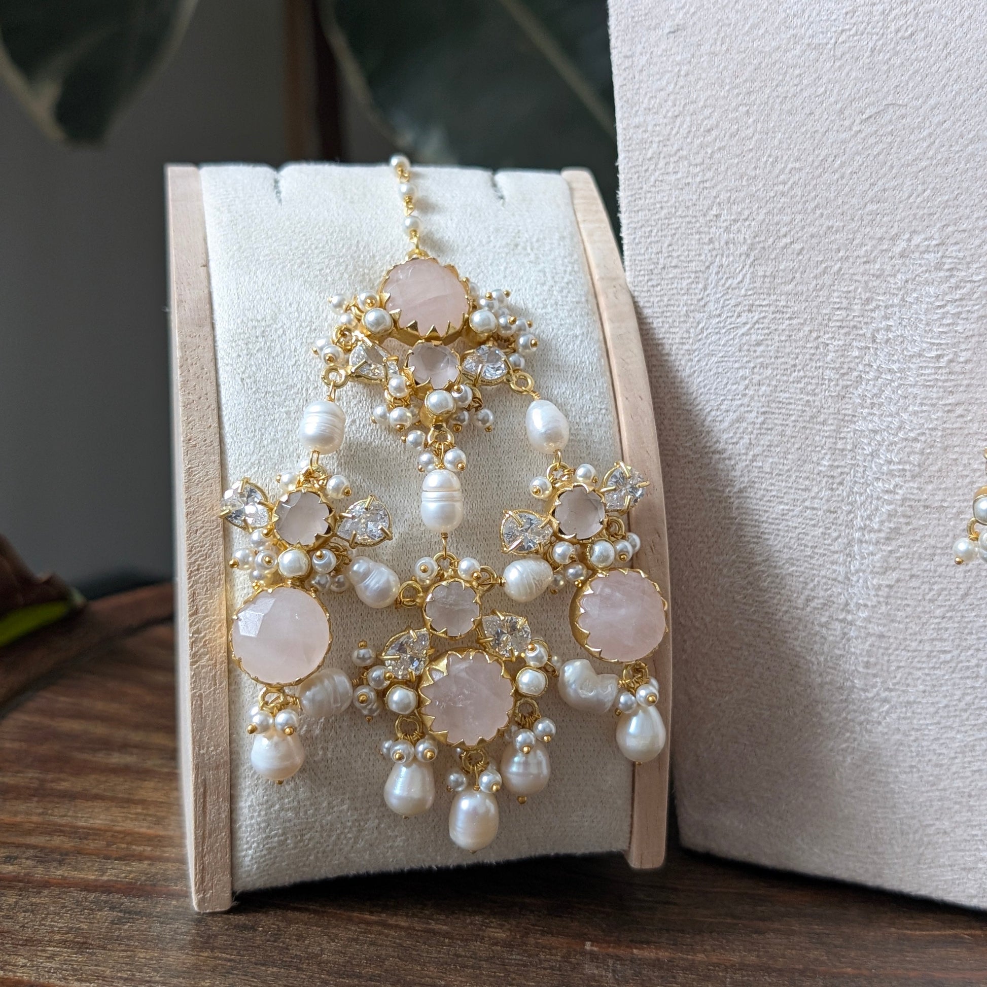 A pair of ornate gold-toned jhumar with white pearls and pale pink stone accents, displayed on a cream jewelry stand on a wooden surface.