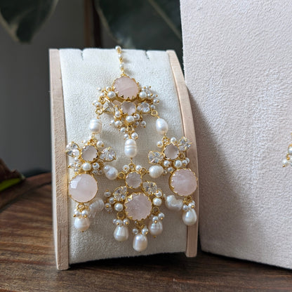 A pair of ornate gold-toned jhumar with white pearls and pale pink stone accents, displayed on a cream jewelry stand on a wooden surface.