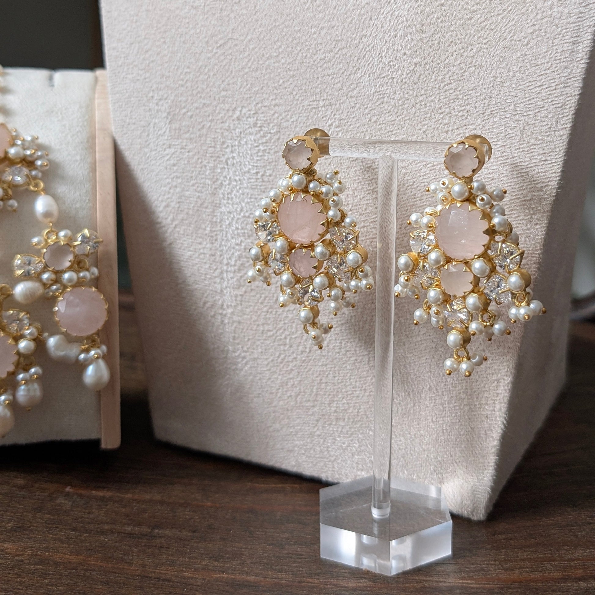 Gold and white pearl chandelier earrings with pale pink stone accents, displayed on a clear stand in front of a cream jewelry bust.