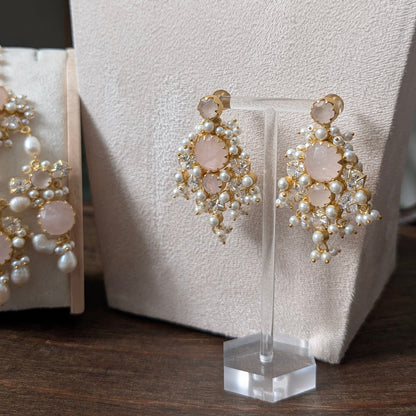 Gold and white pearl chandelier earrings with pale pink stone accents, displayed on a clear stand in front of a cream jewelry bust.
