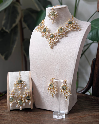 A gold and pearl jewelry set displayed on a cream-colored mannequin bust and a wooden stand, featuring a necklace, matching earrings, and a pair of dangling earrings with green stones, set against a background of indoor plants.