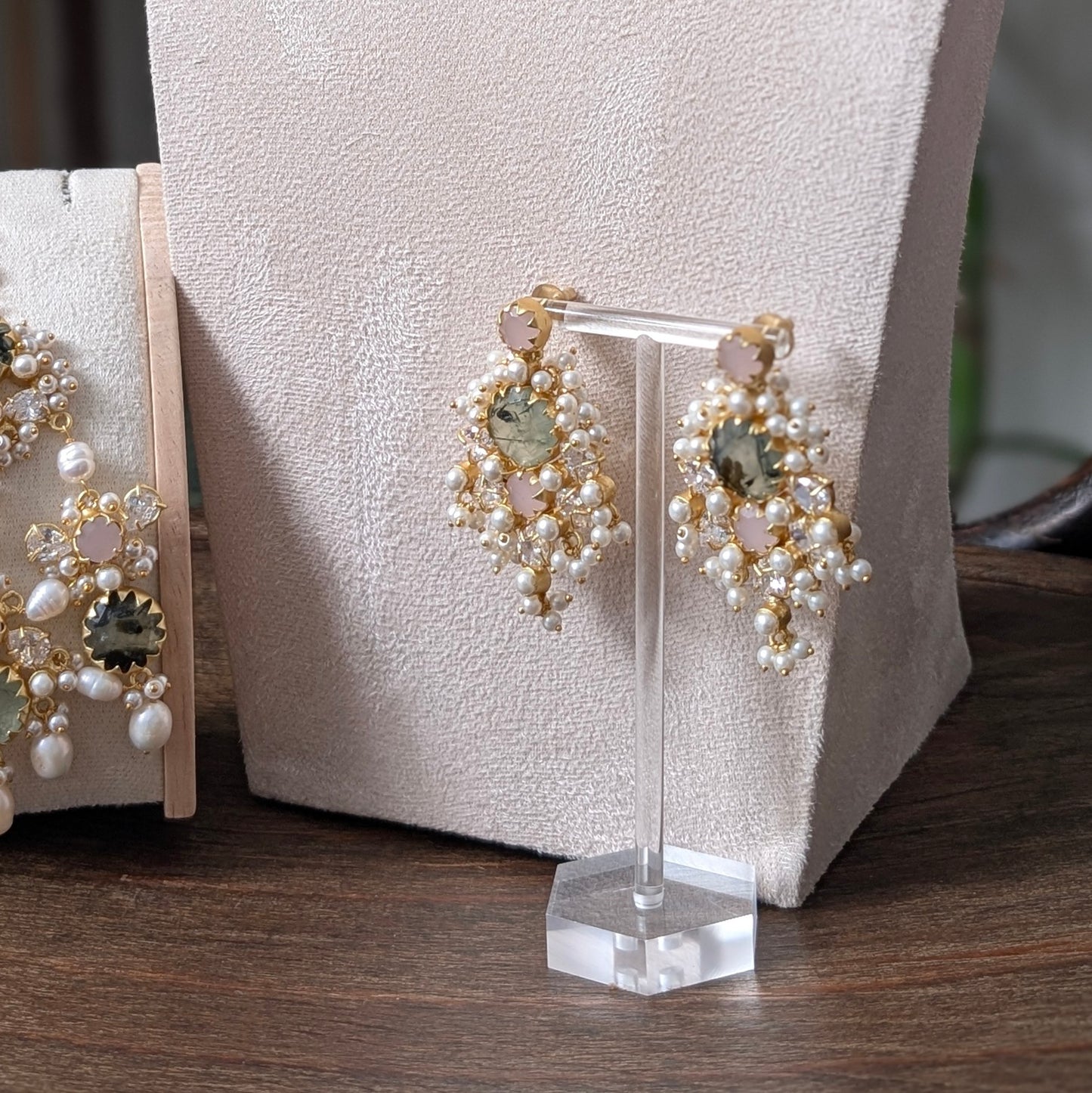 Gold and pearl chandelier drop earrings with green and pink stones, displayed on a clear acrylic stand on a wooden table.