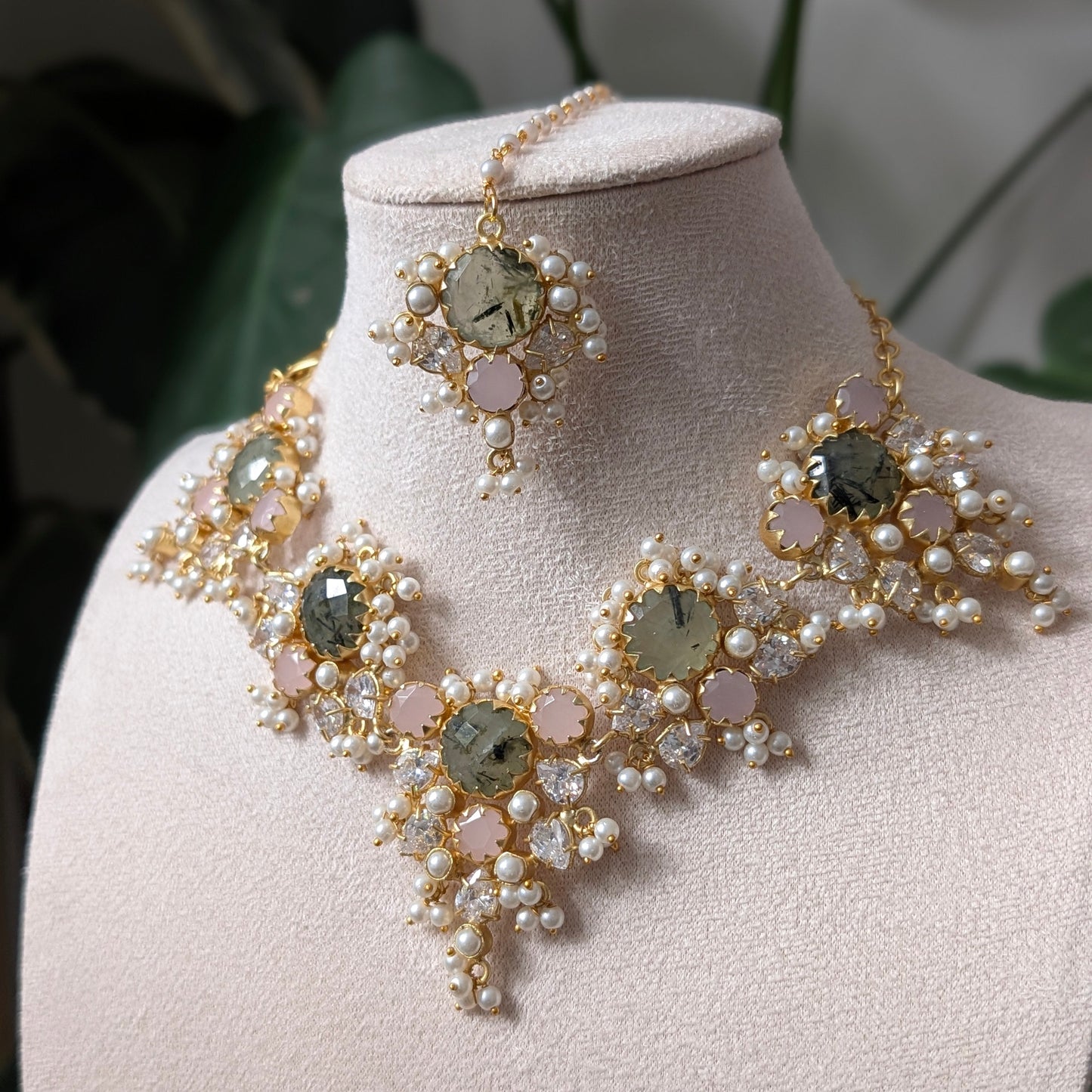 An ornate gold-toned jewelry set displayed on a cream-colored mannequin bust, featuring a statement collar necklace, matching earrings, and a small bracelet. The jewelry is decorated with clusters of white faux pearls, light pink round beads, and large oval iridescent green gemstones.