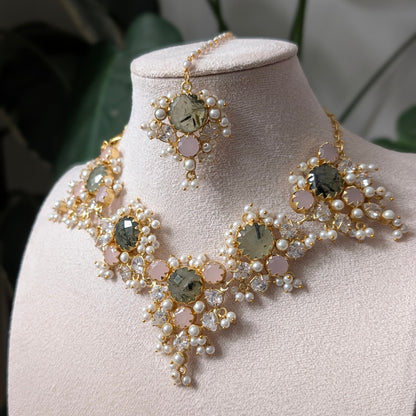 An ornate gold-toned jewelry set displayed on a cream-colored mannequin bust, featuring a statement collar necklace, matching earrings, and a small bracelet. The jewelry is decorated with clusters of white faux pearls, light pink round beads, and large oval iridescent green gemstones.