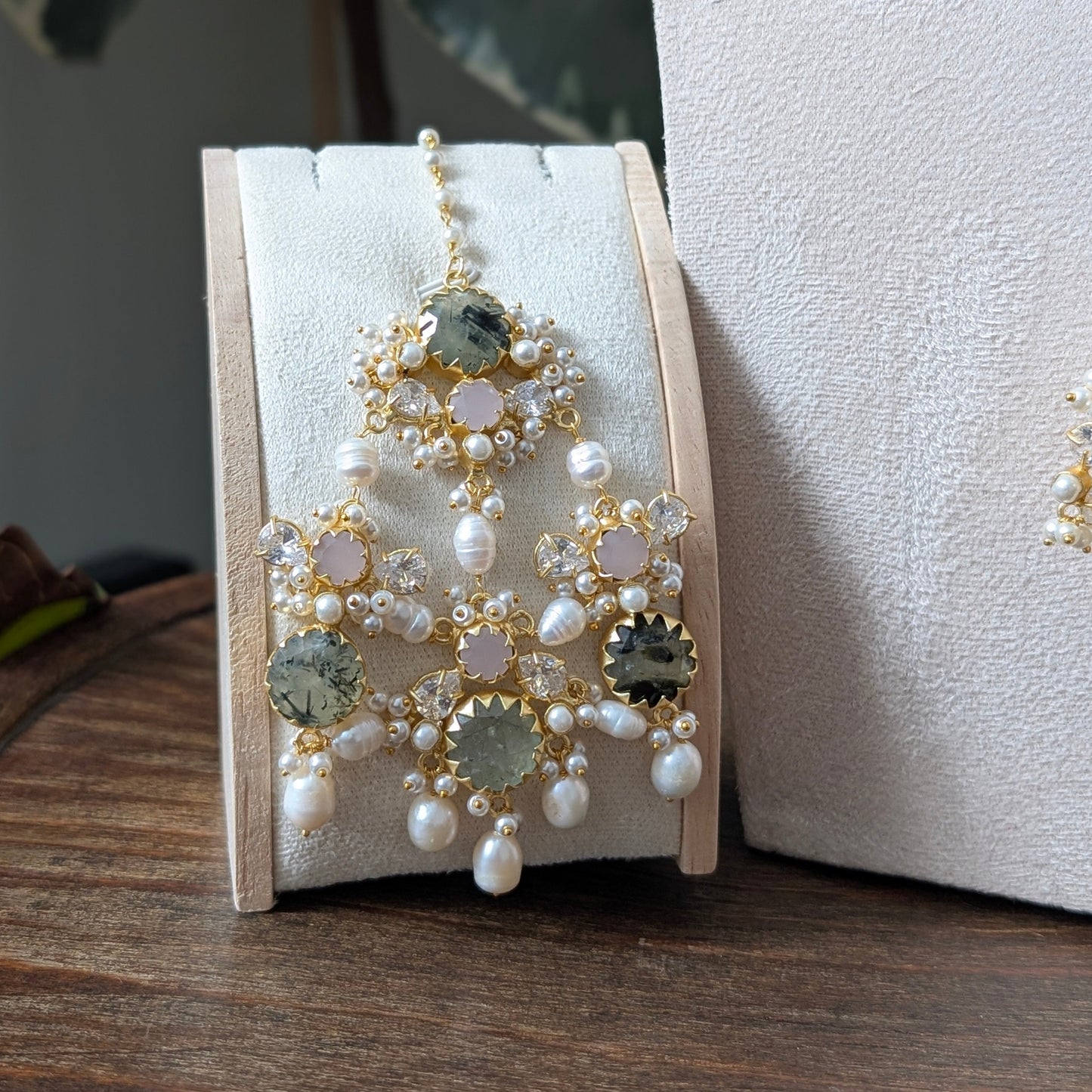 A pair of ornate gold-toned statement jhumar with white pearls and green gemstones, displayed on a cream-colored jewelry stand on a wooden surface.