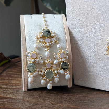 A pair of ornate gold-toned statement jhumar with white pearls and green gemstones, displayed on a cream-colored jewelry stand on a wooden surface.
