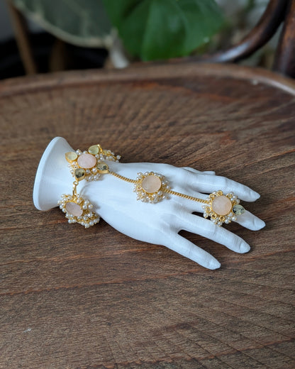 Gold-toned hand chain jewelry with pale pink stone accents and pearl-like details, displayed on a white hand-shaped mannequin over a wooden surface.