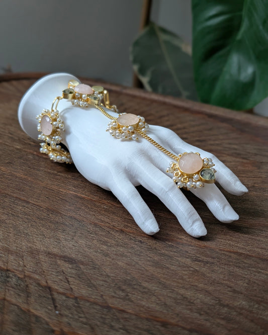 Gold-toned hand chain jewelry with peach-pink stones and pearl-like accents, displayed on a white mannequin hand resting on a wooden surface.