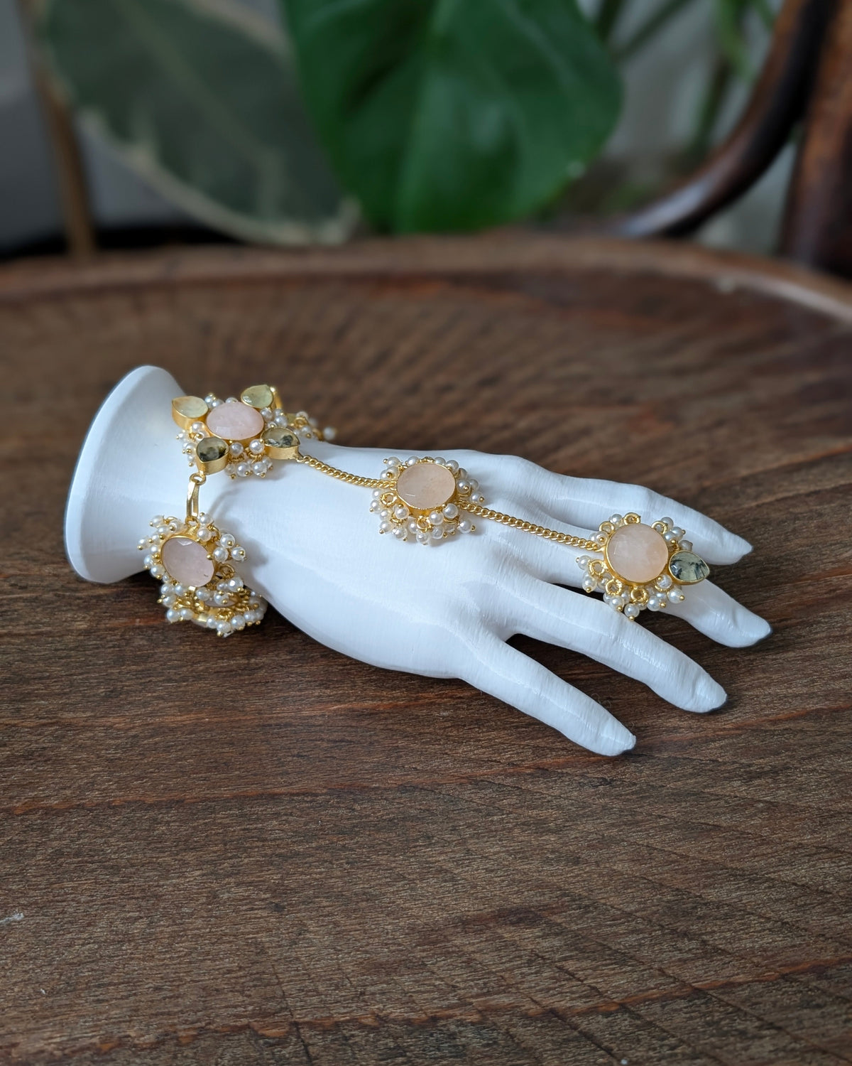 Gold-toned hand chain jewelry with peach-pink stone centers and pearl-like accents, displayed on a white mannequin hand over a wooden surface.