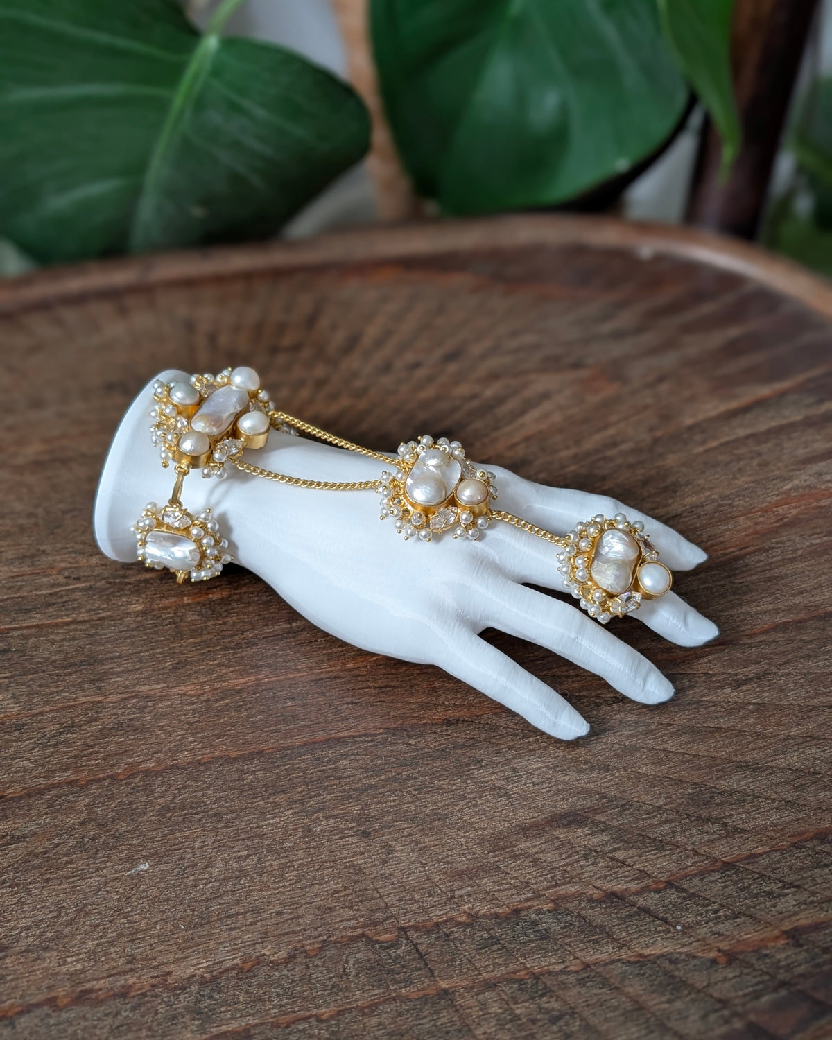 A gold-toned chain hand bracelet with large white pearls and small clear stones, displayed on a white mannequin hand over a rustic wooden surface with green leaves in the background.