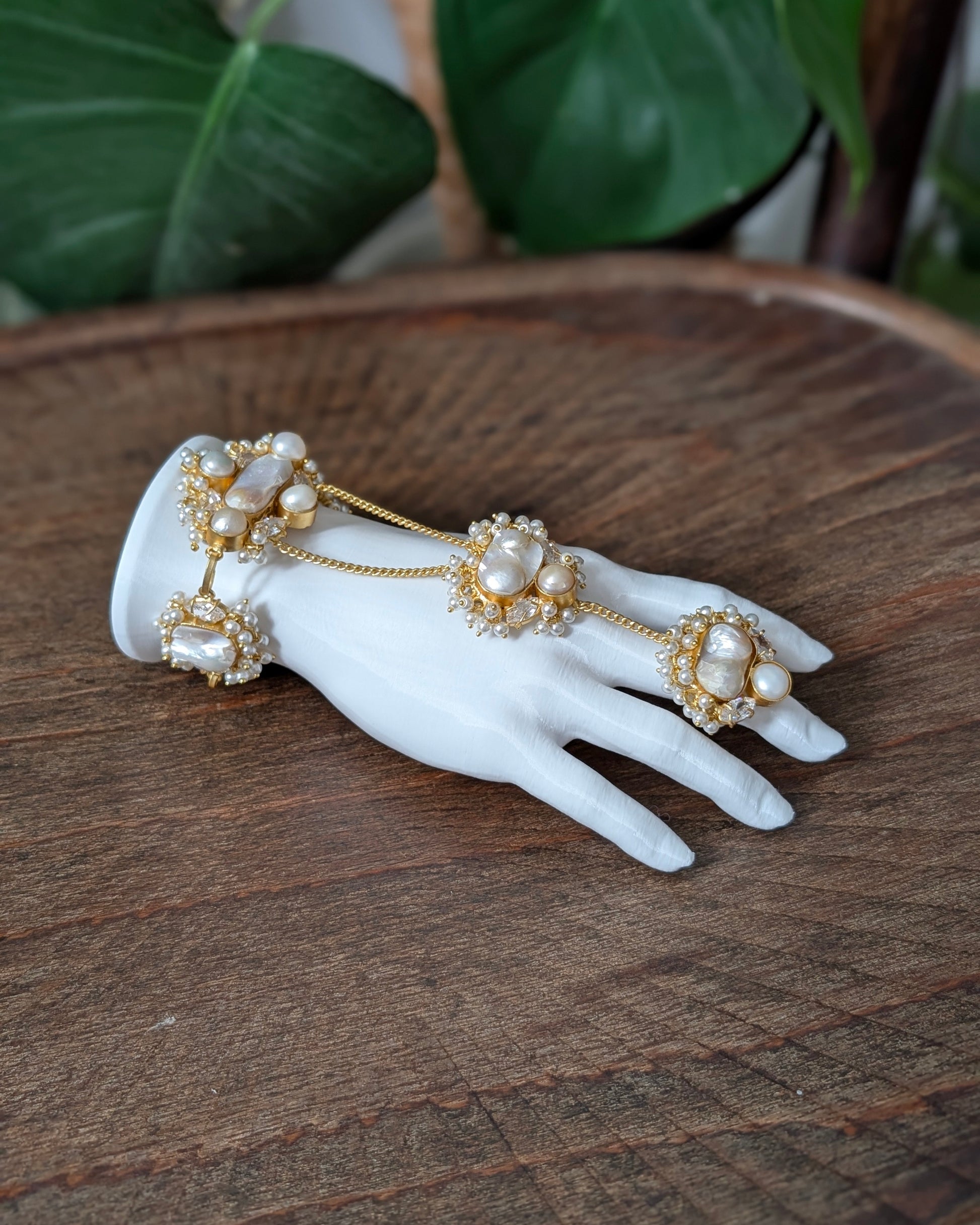 A gold-toned chain hand bracelet with large white pearls and small clear stones, displayed on a white mannequin hand over a rustic wooden surface with green leaves in the background.