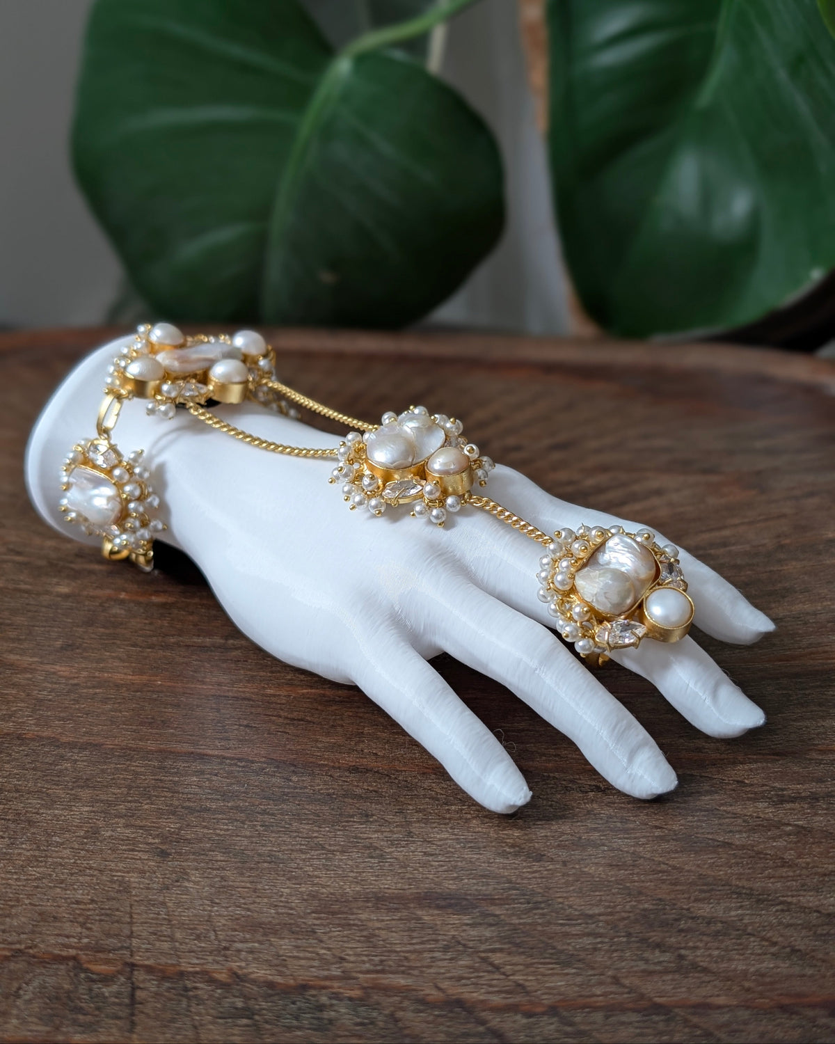 A gold and pearl hand chain jewelry piece displayed on a white mannequin hand, resting on a rustic wooden surface with green leaves in the background.