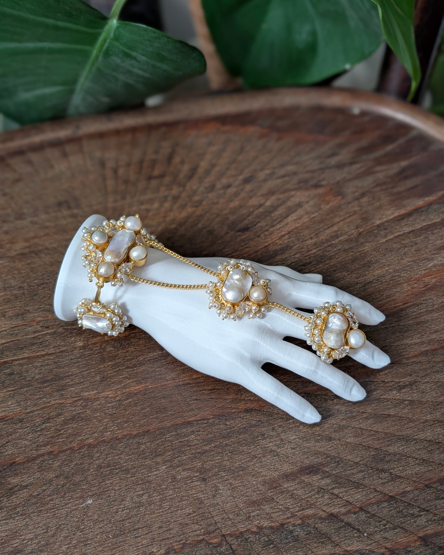 A gold-toned hand chain with pearl-like beads and small clear stones, displayed on a white mannequin hand over a rustic wooden surface with green leaves in the background.