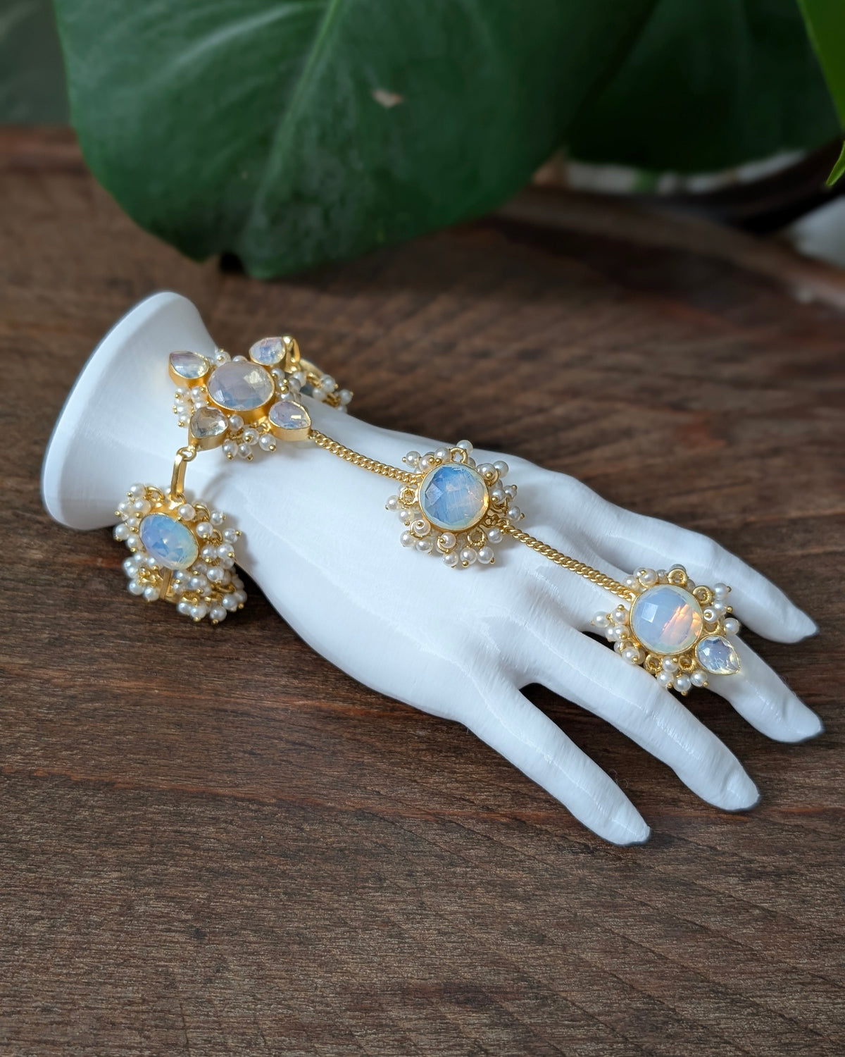 Gold-toned hand chain jewelry with three oval light blue opal-like stones and pearl-like floral accents, displayed on a white hand-shaped mannequin over a wooden surface with green leaves in the background.