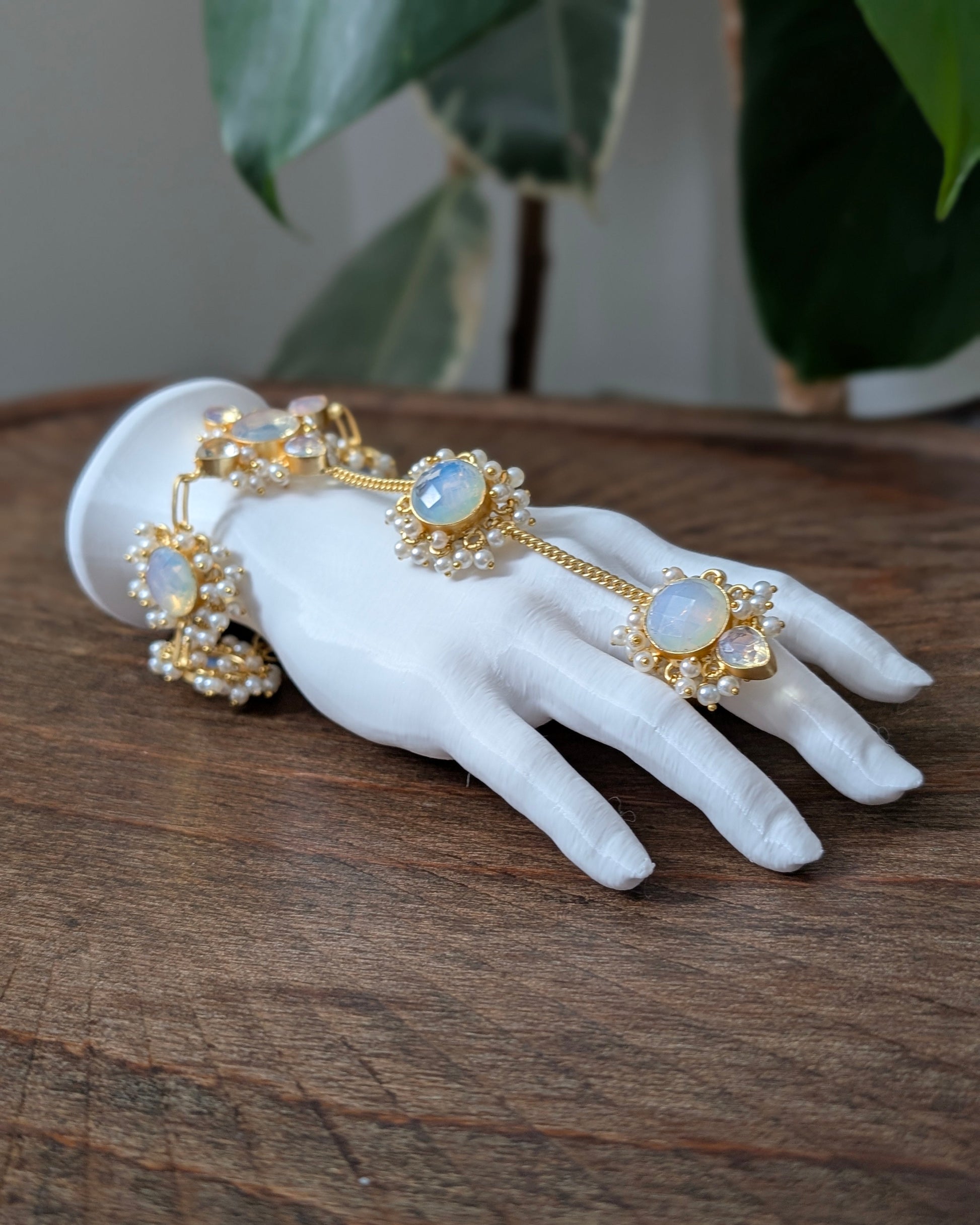 Gold-toned ornate jewelry set with white opal-like stones and pearl-like accents, displayed on a white mannequin hand resting on a wooden surface with blurred green leaves in the background.