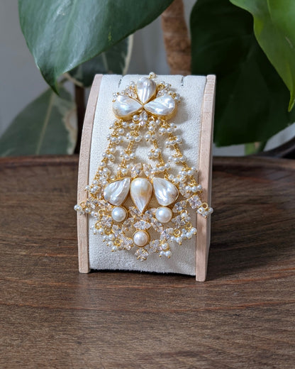 A pearl and gold beaded brooch displayed on a small wooden stand, set on a wooden table with green leaves in the background.