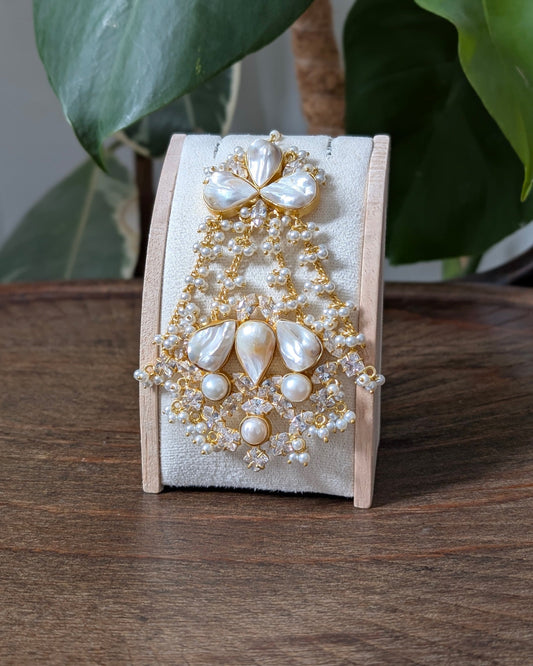 A pearl and gold beaded brooch displayed on a small wooden stand, set on a wooden table with green leaves in the background.
