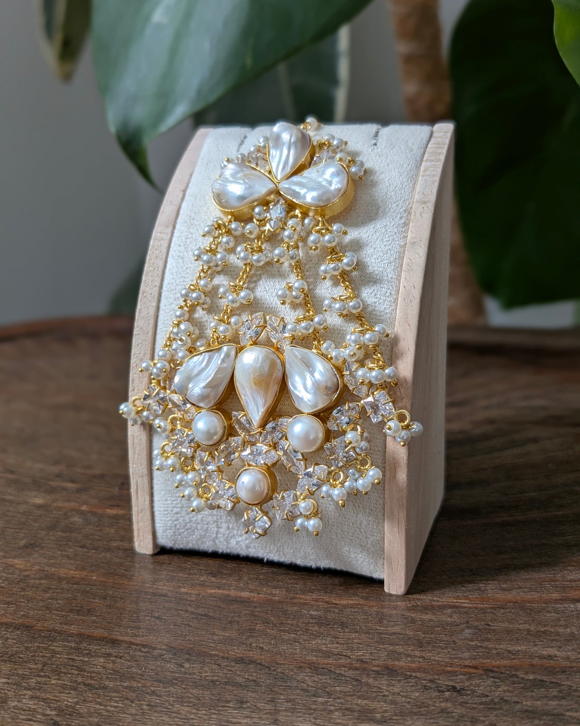 An ornate gold and pearl brooch displayed on a light wood stand on a wooden table, with green leaves in the background.