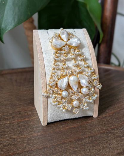 An ornate gold and pearl brooch displayed on a light wood stand on a wooden table, with green leaves and a wooden chair visible in the background.
