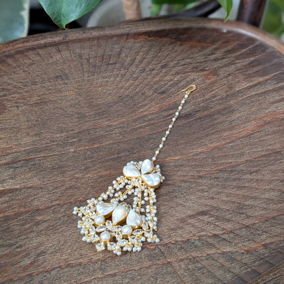 A gold and white beaded tassel earring with large white pearl-like drops, displayed on a rustic wooden tray with green leaves in the background.