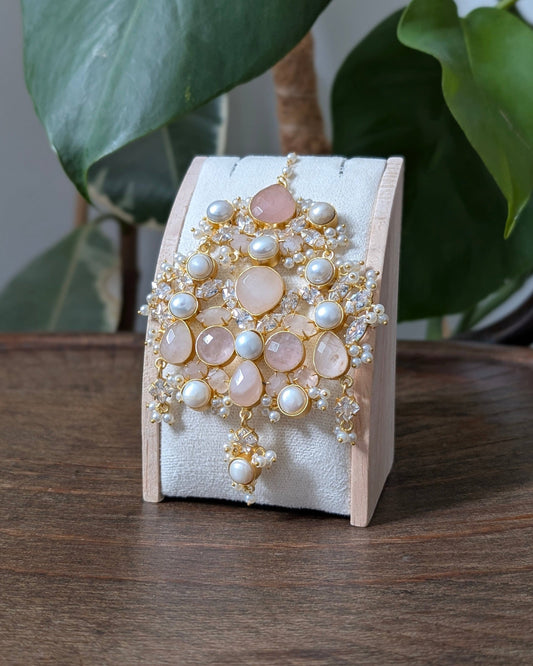 A gold-toned statement brooch featuring pale pink stones and white faux pearls, displayed on a white fabric jewelry stand on a wooden table with green plants in the background.